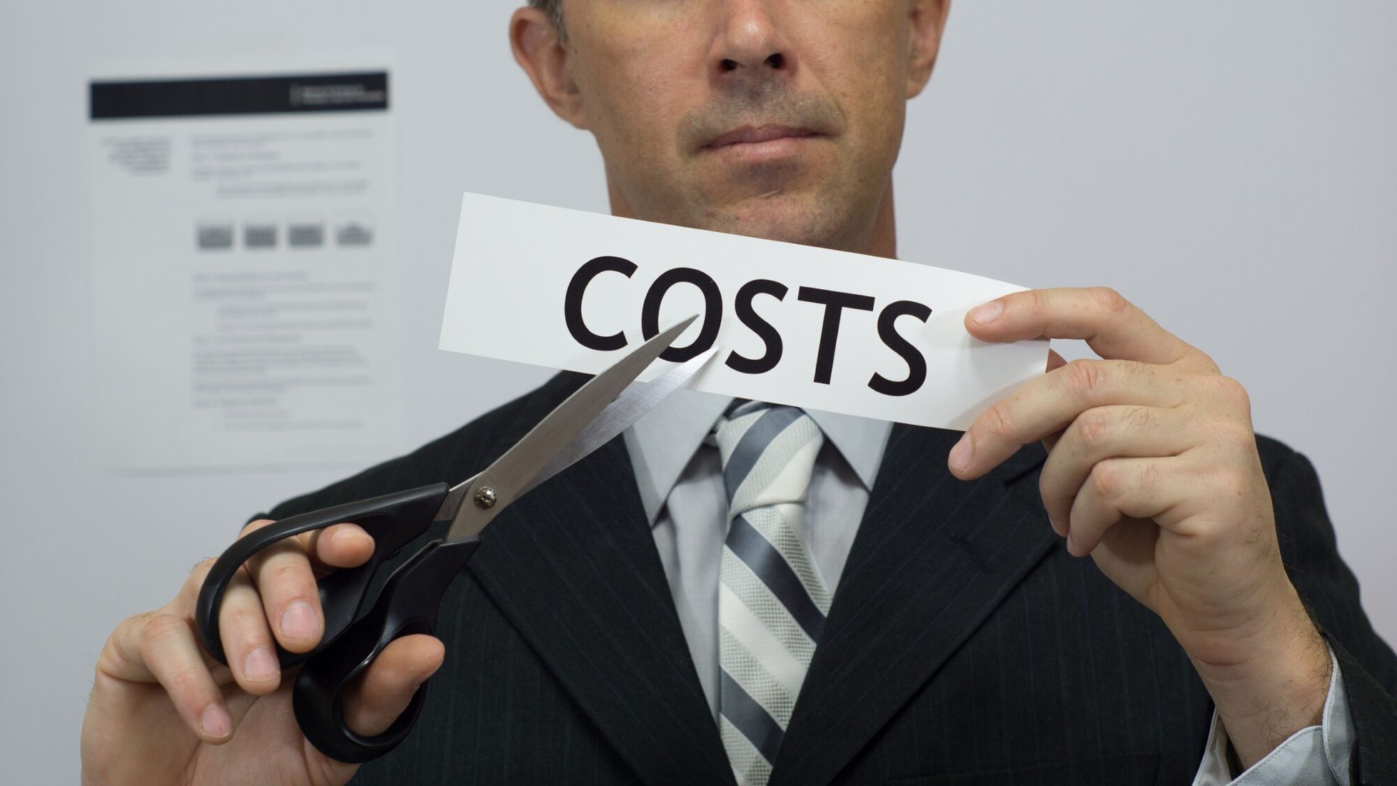 A business person in a suit and tie holds scissors and cuts a piece of paper with the word COSTS.